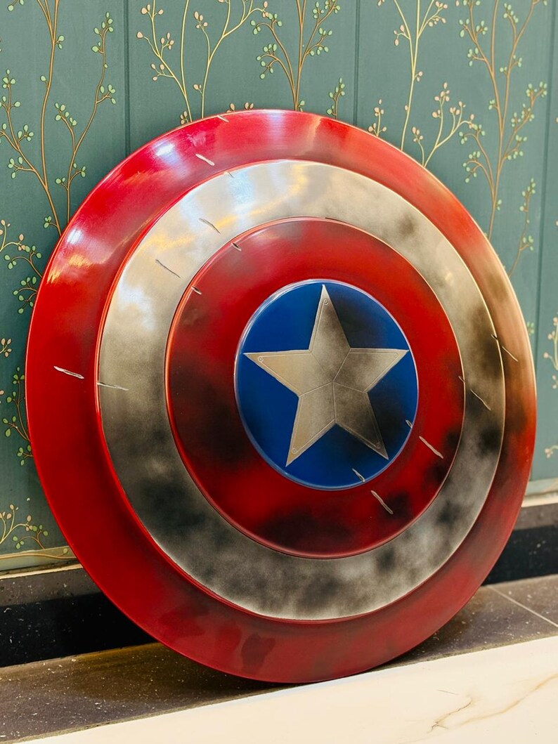 Puede incluir: Un escudo redondo con una combinaci&oacute;n de colores rojo, blanco y azul. El escudo presenta un c&iacute;rculo azul central con una estrella plateada, rodeado de anillos conc&eacute;ntricos rojos y plateados. El escudo tiene un aspecto desgastado.