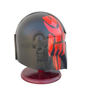 May include: Black helmet with red accents, displayed on a wooden stand. The helmet features a distinctive red design on the front and top. The helmet is a replica of a character's helmet from a science fiction franchise.