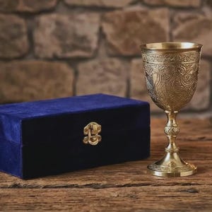 May include: A gold-colored metal chalice with intricate embossed floral patterns on the bowl and stem, positioned next to a rectangular blue velvet storage box featuring a central gold-toned metal latch closure on the front panel.