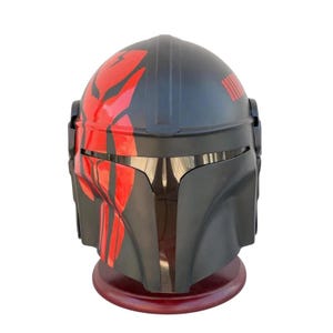 May include: A black and red Mandalorian helmet with a glossy finish. The helmet features red markings on the top and side, with a visor and a series of lines on the back. It is displayed on a dark wooden stand.