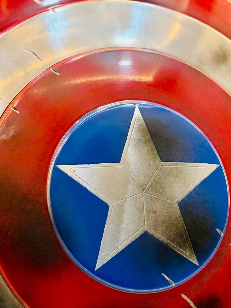 Puede incluir: Primer plano de un escudo con una estrella plateada sobre un c&iacute;rculo azul, sobre un fondo rojo y plateado. El escudo tiene un dise&ntilde;o circular con un acabado met&aacute;lico, que recuerda el emblema de un superh&eacute;roe.