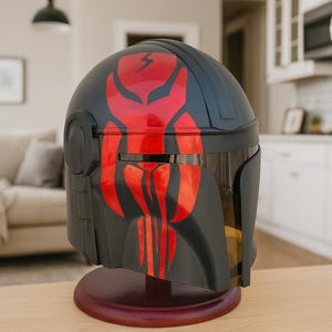 May include: A black and red helmet with a distinctive design, resting on a wooden stand. The helmet features a red emblem on the front and top, with a visor. The design suggests a futuristic or science fiction theme.