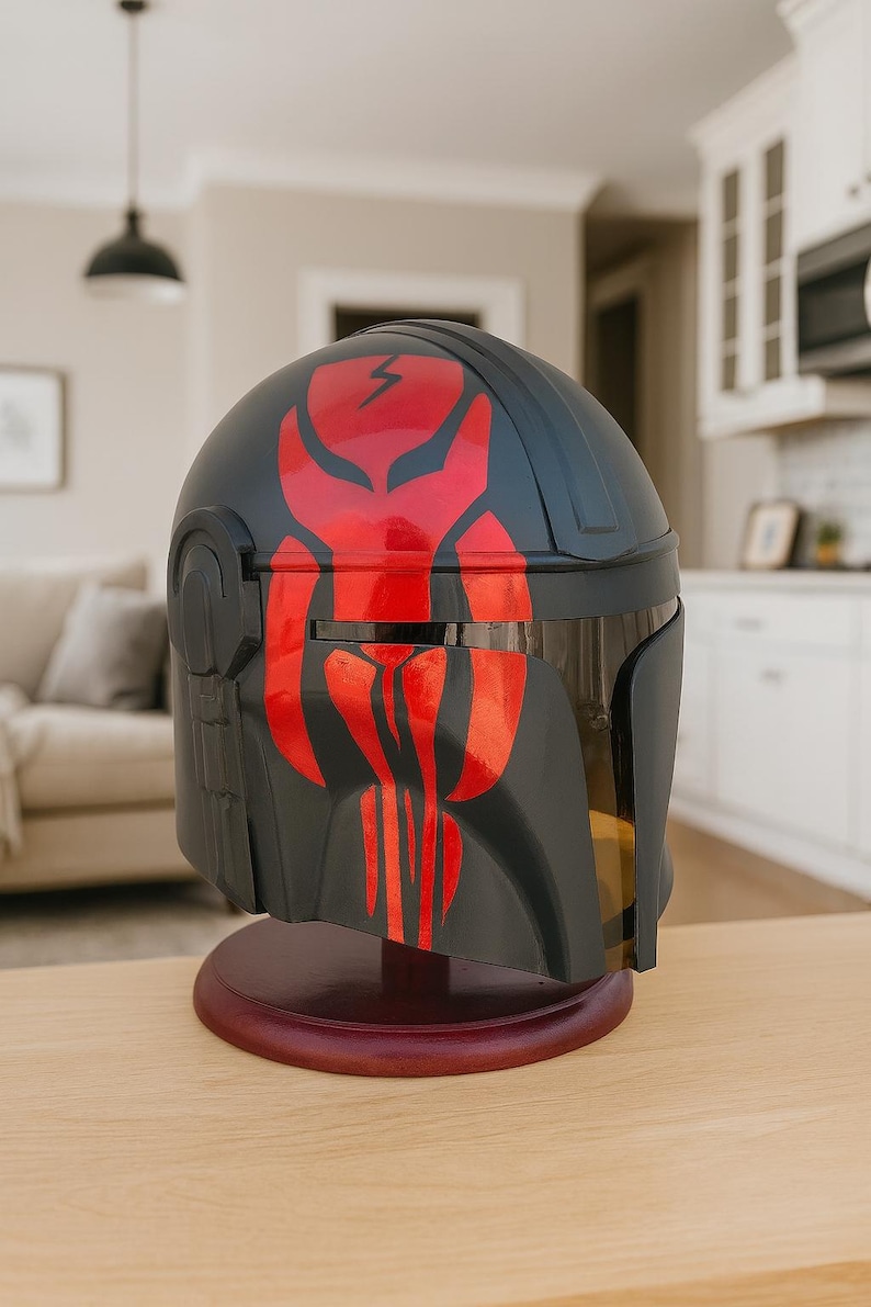 May include: A black and red helmet with a distinctive design, resting on a wooden stand. The helmet features a red emblem on the front and top, with a visor. The design suggests a futuristic or science fiction theme.