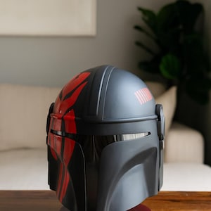 May include: A black and red Mandalorian helmet, displayed on a wooden stand. The helmet features red markings and a visor. The helmet is a replica of the one worn by the character in the Star Wars series.