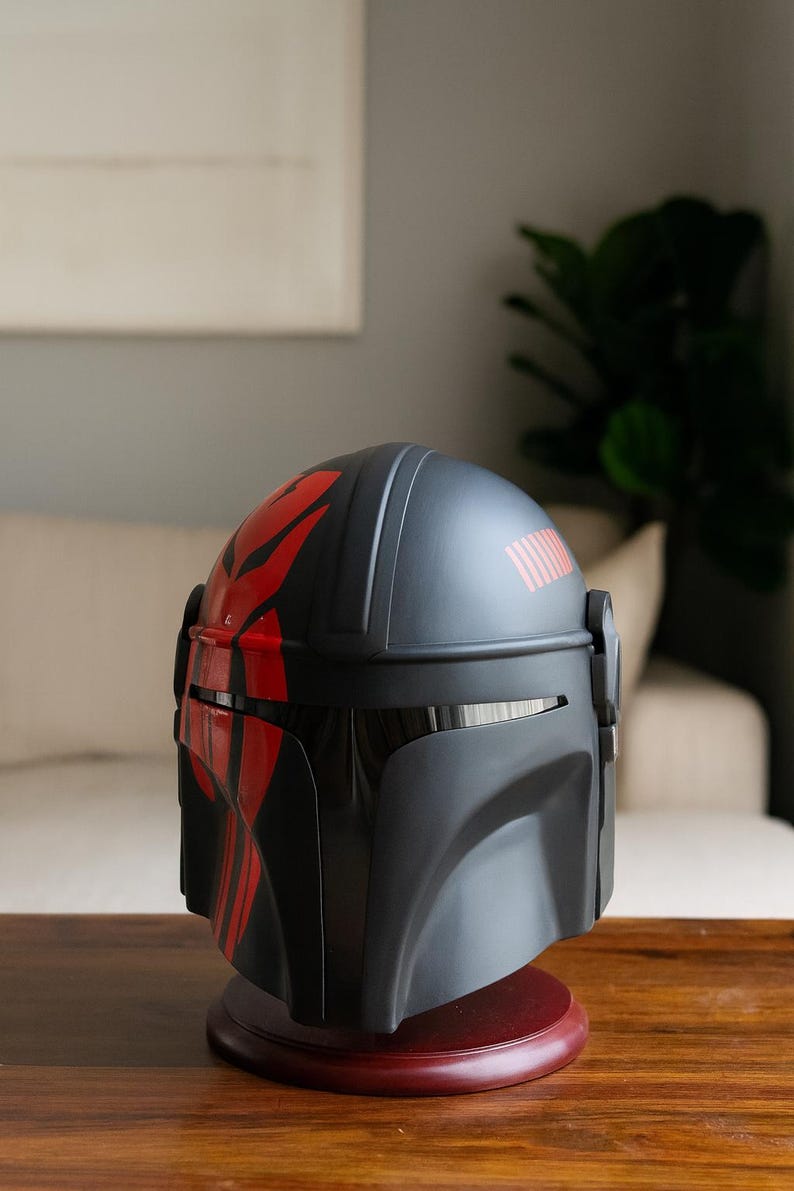 May include: A black and red Mandalorian helmet, displayed on a wooden stand. The helmet features red markings and a visor. The helmet is a replica of the one worn by the character in the Star Wars series.
