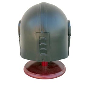 May include: A dark green helmet with a rounded top and vertical ridges. The helmet has side details and a series of rectangular features on the back. It is displayed on a round, reddish-brown stand.