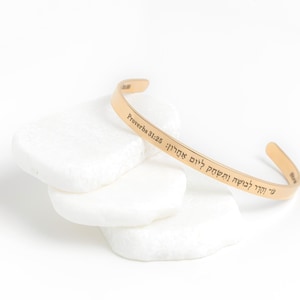 May include: Gold bangle bracelet with the inscription "Proverbs 31:25" in Hebrew. The bracelet is on a white background.