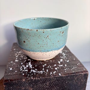 May include: A handmade ceramic bowl with a speckled turquoise glaze on the upper half and a cream-colored base. The bowl is set on a dark wooden surface, showcasing its rustic charm. This piece is ideal for serving food or as a decorative item.