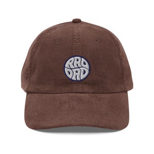 Puede incluir: Una gorra de béisbol de pana marrón con un círculo bordado blanco que contiene el texto "RAD DAD" en una fuente retro.