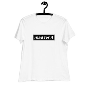 Puede incluir: Camiseta blanca con el texto "mad fer it" en un rectángulo negro. La camiseta está colgada de una percha negra. El texto está en negrita, con una fuente sans-serif.