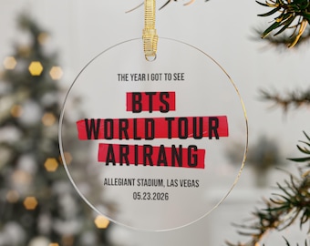 May include: A circular clear acrylic ornament hanging from a gold ribbon. The text reads: THE YEAR I GOT TO SEE BTS WORLD TOUR ARIRANG, ALLEGIANT STADIUM, LAS VEGAS, 05.23.2026. The words BTS, WORLD TOUR, and ARIRANG are highlighted by red brush strokes.