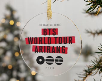 May include: A clear, circular acrylic ornament hanging from a gold ribbon. The text reads THE YEAR I GOT TO SEE BTS WORLD TOUR ARIRANG 2026. Below the text are three circular icons featuring a solid dot, horizontal lines, and a segmented bar.