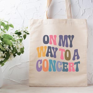 Puede incluir: Bolso tote de lona beige con el texto "ON MY WAY TO A CONCERT" en letras coloridas de estilo retro. El bolso tiene asas largas y se muestra sobre un fondo blanco texturizado. Un jarrón con flores blancas y hojas verdes está a la izquierda.