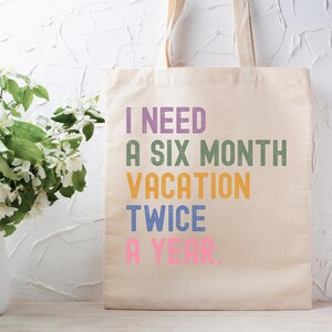 May include: A beige canvas tote bag featuring the text "I NEED A SIX MONTH VACATION TWICE A YEAR" in purple, green, orange, blue, and pink. The bag has two beige handles. The background is white and textured.