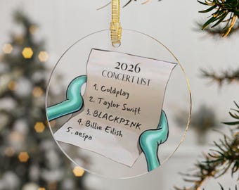 May include: A circular clear acrylic ornament featuring a printed list titled 2026 CONCERT LIST with five entries: 1. Coldplay, 2. Taylor Swift, 3. BLACKPINK, 4. Billie Eilish, 5. aespa, held by two teal cartoon arms, suspended by a gold ribbon.