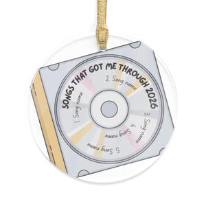 May include: A circular ornament featuring a graphic of a compact disc inside a square case. The text reads SONGS THAT GOT ME THROUGH 2026, with numbered slots for five song names. The design includes light gray, tan, and pink accents with a gold ribbon.