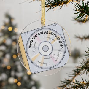 May include: A circular clear acrylic ornament featuring a printed graphic of a compact disc case. The design includes the text "SONGS THAT GOT ME THROUGH 2026" and five numbered slots labeled "Song name" arranged around the center disc graphic.