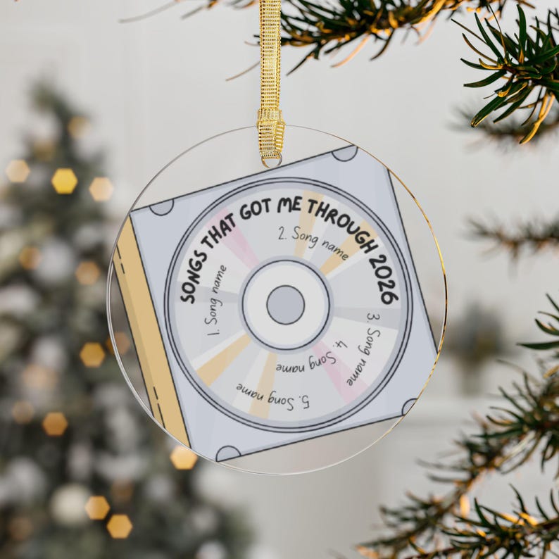 May include: A circular clear acrylic ornament featuring a printed graphic of a compact disc case. The design includes the text "SONGS THAT GOT ME THROUGH 2026" and five numbered slots labeled "Song name" arranged around the center disc graphic.