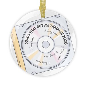 May include: A round white ceramic ornament featuring a graphic of a compact disc inside a case. The text reads SONGS THAT GOT ME THROUGH 2026 with five numbered slots for song names. It includes a gold ribbon loop for hanging at the top.