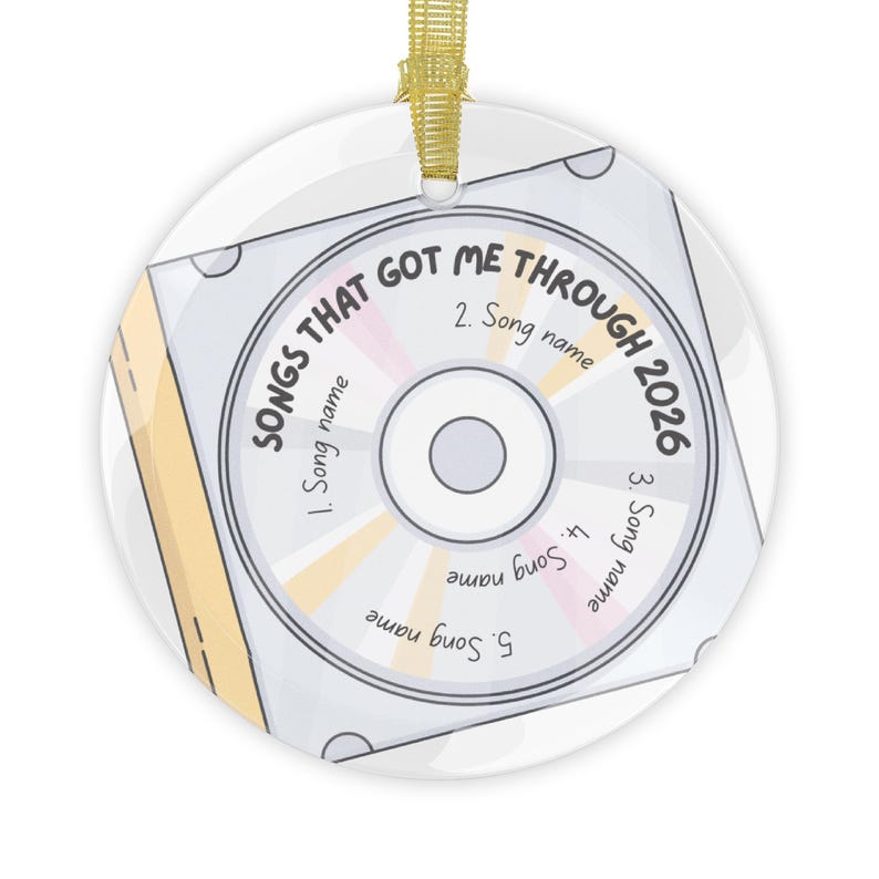 May include: A round white ceramic ornament featuring a graphic of a compact disc inside a case. The text reads SONGS THAT GOT ME THROUGH 2026 with five numbered slots for song names. It includes a gold ribbon loop for hanging at the top.