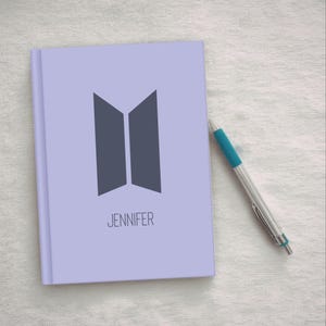May include: A lavender notebook with a dark gray geometric design and the name "JENNIFER" printed below. A silver and blue pen rests beside the notebook on a white surface.