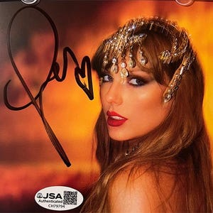 May include: A signed photograph featuring a woman with red lipstick and a jeweled headpiece. The autograph is in black ink. The image has a warm, orange and yellow background. A JSA authentication sticker is in the lower left corner.