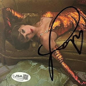 May include: An image of a woman wearing a sequined orange dress, lying on a green couch. The image is signed with a black marker. A JSA Authenticated sticker is in the lower left corner.