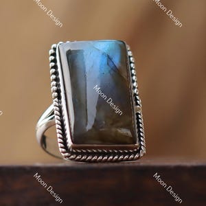 Puede incluir: Anillo rectangular de plata con una gran piedra de labradorita iridiscente. La piedra muestra tonos azules y grises. El anillo tiene un borde detallado en forma de cuerda. El anillo está sobre una superficie de madera oscura.