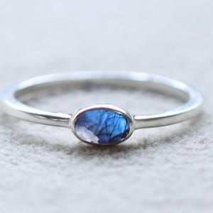Puede incluir: Un anillo de plata con una pequeña gema azul de forma ovalada. La banda del anillo es delgada y delicada, y la gema está engastada en un bisel simple. La gema tiene una mezcla de tonos azules y un brillo sutil.