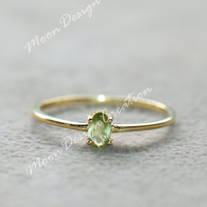 Puede incluir: Un delicado anillo de oro con una pequeña gema ovalada de color verde claro. El anillo tiene una banda fina y un diseño simple y elegante. La gema mide aproximadamente 0,6 cm de largo.