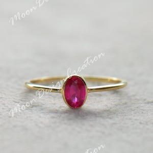 May include: A gold ring with a vivid oval-cut ruby gemstone. The ring has a slender band and a simple, elegant design. The ruby is a deep, rich red colour, set in a minimalist style.