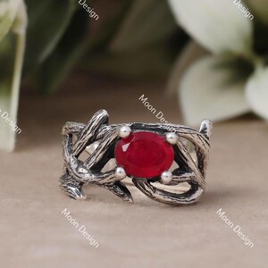 Puede incluir: Un anillo de plata con una gran gema roja de forma ovalada. El anillo tiene un diseño texturizado único que se asemeja a ramas entrelazadas. El anillo está sobre un fondo neutro con follaje verde y blanco.