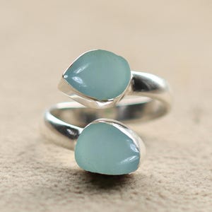 Puede incluir: Un anillo de plata con dos piedras en forma de lágrima de color azul claro. La banda del anillo se curva alrededor del dedo, con cada piedra engastada en un bisel de plata. Las piedras tienen un aspecto liso y pulido.