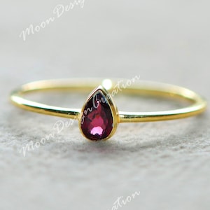 Op de afbeelding: Een delicate gouden ring met een druppelvormige, dieprode edelsteen. De ring heeft een dunne, gepolijste band en een eenvoudig, elegant ontwerp. De edelsteen is gezet in een bezel-zetting.