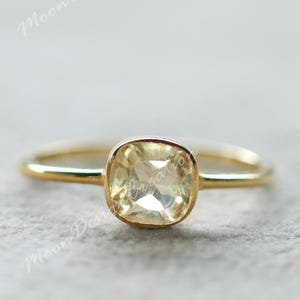 Könnte beinhalten: Ein goldener Ring mit einem quadratischen, hellgelben Edelstein. Der Edelstein ist in einer Zargenfassung gefasst, und das Band ist dünn und schlicht. Der Ring wird auf einer hellgrauen Oberfläche präsentiert.