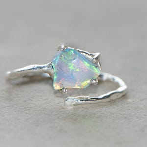 May include: A silver ring with a raw, iridescent opal gemstone. The opal displays a play of colours, including blues, greens, and yellows. The ring's band has a textured, organic design, adding to its unique aesthetic. The ring is set against a neutral background.