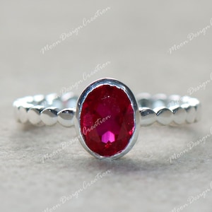 Könnte beinhalten: Ein silberner Ring mit einem großen, oval geschliffenen, leuchtend roten Edelstein. Das Band hat ein zartes, muschelförmiges Design. Der Ring ist vor einem neutralen Hintergrund platziert, um die Farbe des Edelsteins hervorzuheben.