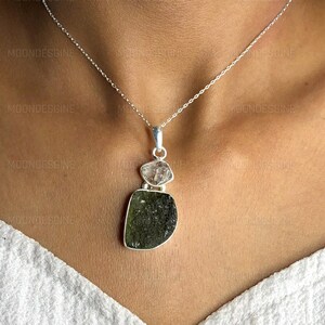 May include: A silver chain necklace with a pendant featuring a layered green and clear crystal. The pendant is made of three pieces of crystal, with the largest piece being a dark green, faceted crystal. The smaller pieces are clear and faceted.