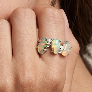 Op de afbeelding: Een zilveren ring met drie ruwe opaalstenen. De opalen tonen een mix van kleuren, waaronder groen, geel en oranje, in een modern, minimalistisch ontwerp. De ring wordt aan een vinger getoond.