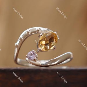 Peut inclure: Une bague en argent ornée d'une grande pierre précieuse jaune doré de forme ovale et d'une petite pierre précieuse violet clair. La bague présente un design tourbillonnant unique avec un anneau texturé. Fond en bois flou.