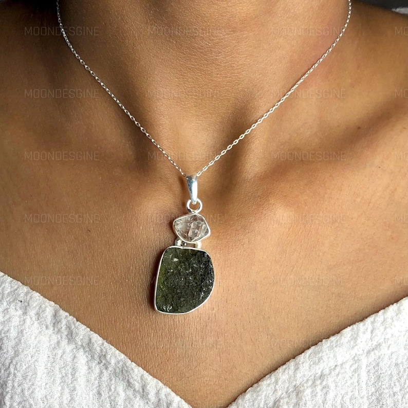 May include: A silver chain necklace with a pendant featuring a layered green and clear crystal. The pendant is made of three pieces of crystal, with the largest piece being a dark green, faceted crystal. The smaller pieces are clear and faceted.