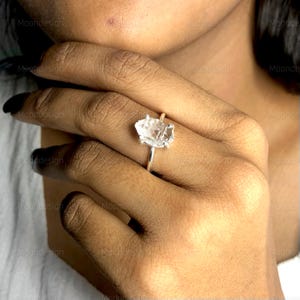 Peut inclure: Une bague en argent avec une pierre précieuse en cristal brut clair et facetté. La bague est portée au doigt d'une personne.