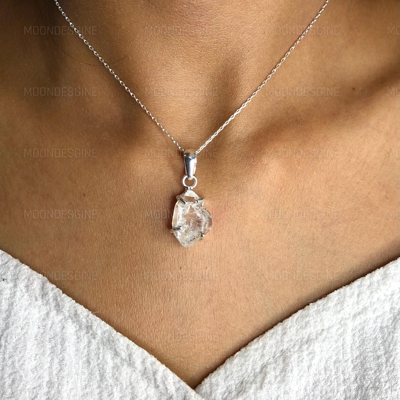 Puede incluir: Un collar de cadena de plata con un colgante de cristal de cuarzo transparente. El cristal es en bruto y tiene una forma natural irregular.