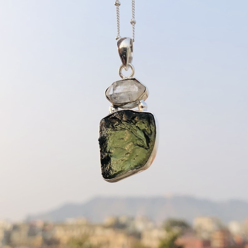 May include: A silver chain necklace with a pendant featuring a green and black gemstone and a clear crystal.