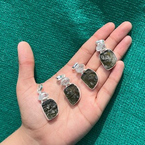 May include: Four silver pendants featuring green Moldavite and clear quartz crystals. The pendants are shown on a hand against a green backdrop.