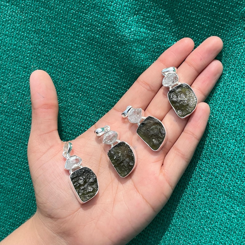 May include: Four silver pendants featuring green Moldavite and clear quartz crystals. The pendants are shown on a hand against a green backdrop.
