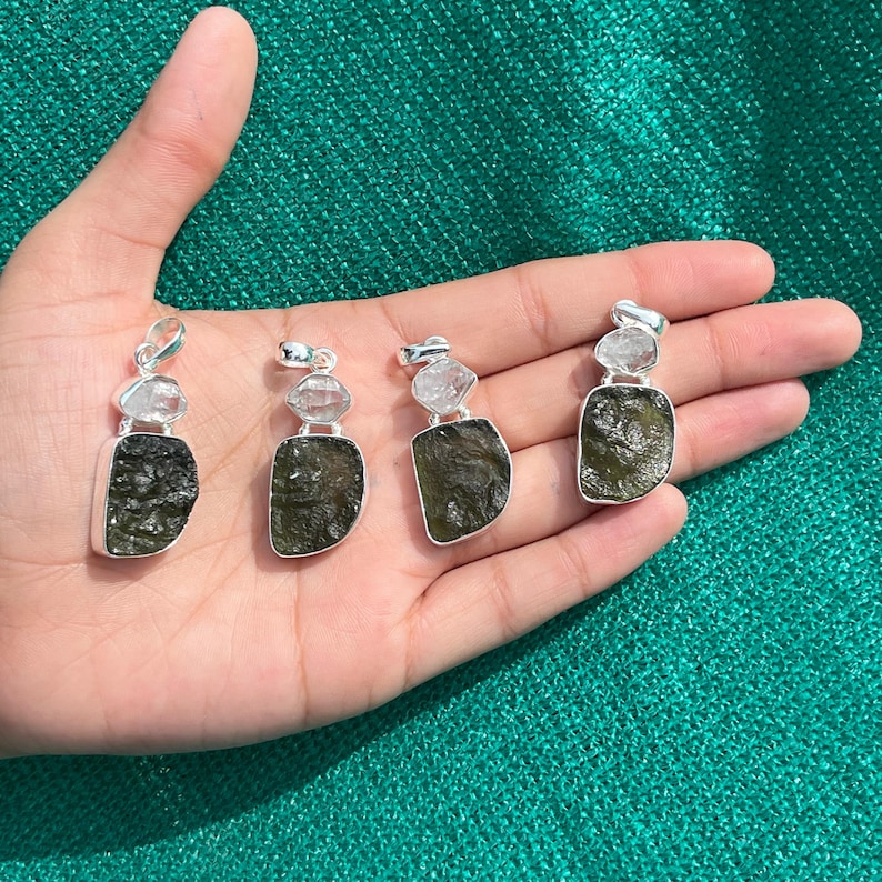 May include: Four silver pendants featuring green and clear stones. Each pendant has a green stone with a clear stone on top. The pendants are shown on a hand.
