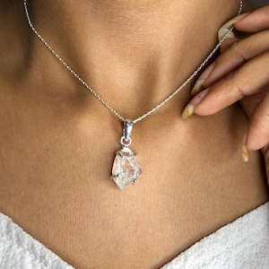Puede incluir: Un collar de cadena de plata con un colgante de cristal de cuarzo transparente. El cristal est&aacute; facetado y tiene un aspecto &aacute;spero y natural.