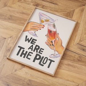 May include: A white framed print with the text "We are the plot" in black lettering. The print features a watercolor illustration of two hands holding a martini glass and a champagne flute.