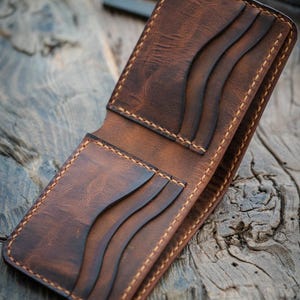 Puede incluir: Una cartera de cuero marrón con múltiples ranuras para tarjetas y un portafolios. La cartera tiene un aspecto desgastado y está cosida con hilo marrón claro.